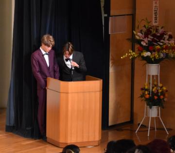 花輪が設置されたステージ横の演台の前でスーツ姿の男性2名が立っておりそのうち1名がマイクを持ち話をしている写真