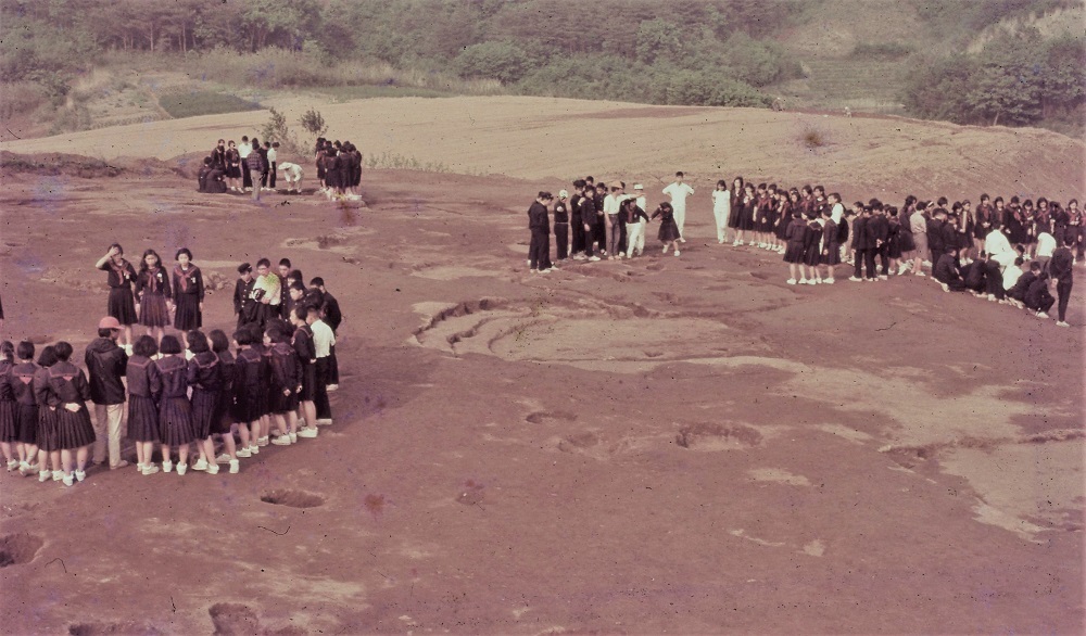 6号住居跡付近で大勢の中学生がグループに分かれて作業員に説明を聞きながら見学をしている写真