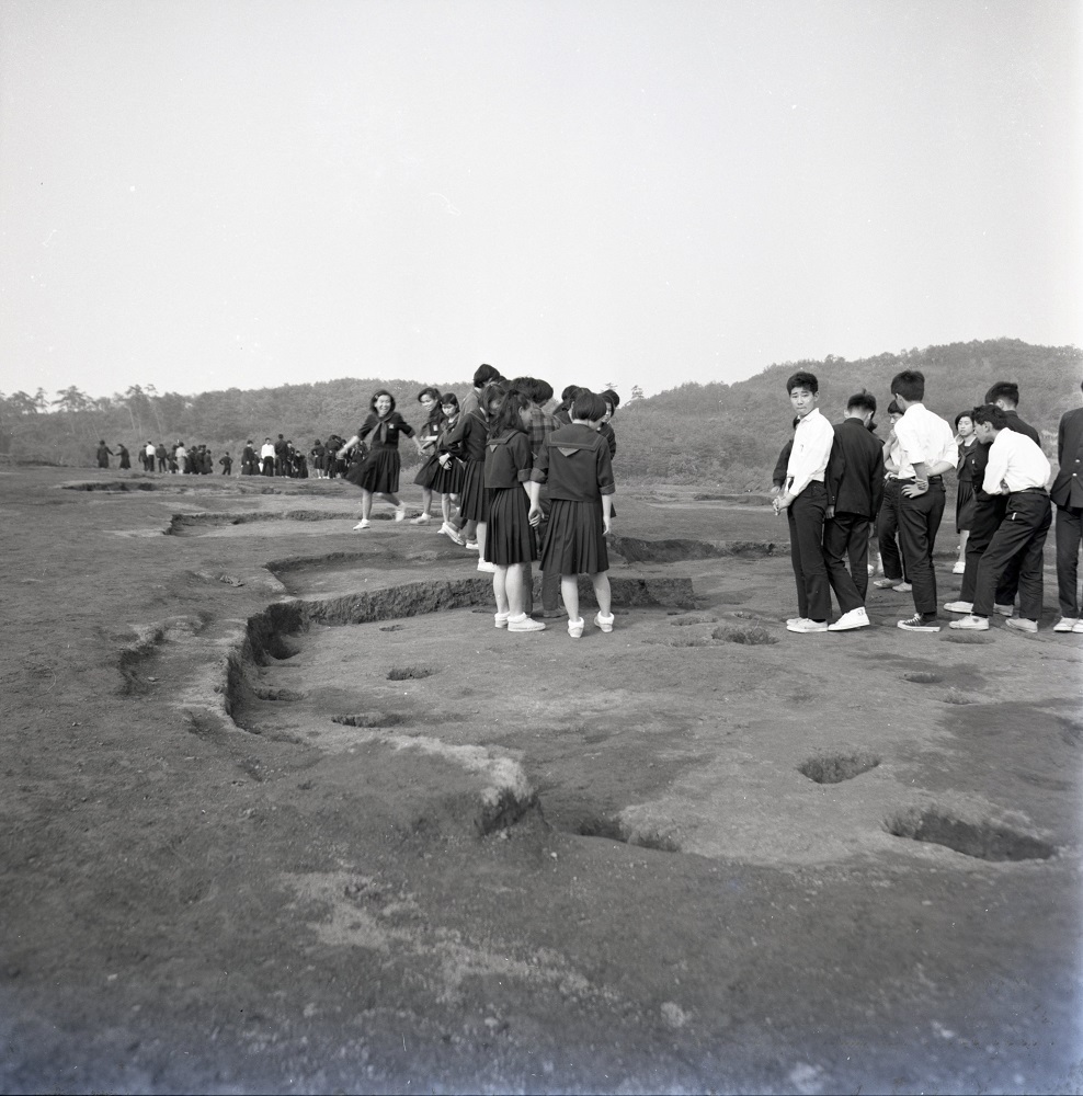 溝や穴が掘られている11号住居跡付近で中学生たちが見学を行っている白黒写真