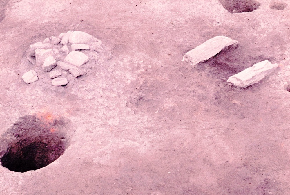 左上に両手のひらほどの石が積み上げられ右上に長方形の石が2つ横に並べられており左下に円形の穴が掘られている住居跡の写真