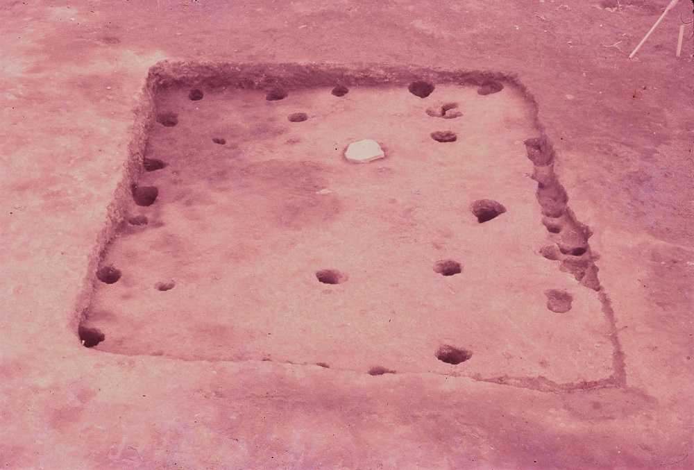 正方形に掘られた溝の中に複数の穴が掘られており、中心あたりに石が1つだけ置かれている住居跡の写真