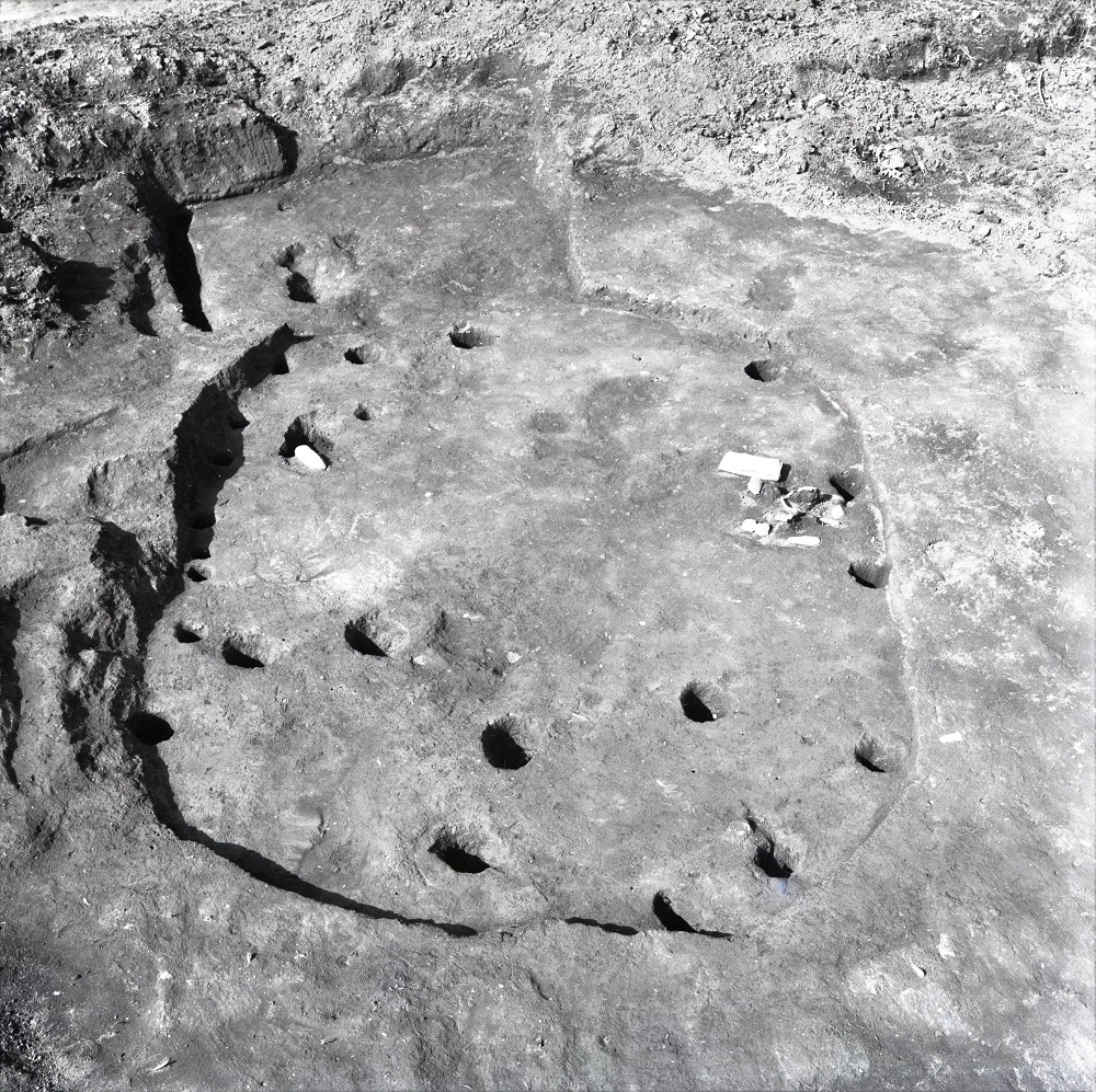 円形に掘られた溝の内側に複数の穴や石が点在している住居跡の写真
