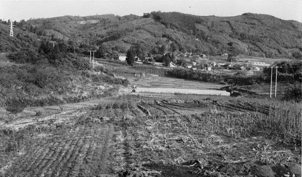 後方に山々や住宅街があり手前に田畑が広がっている白黒の風景写真