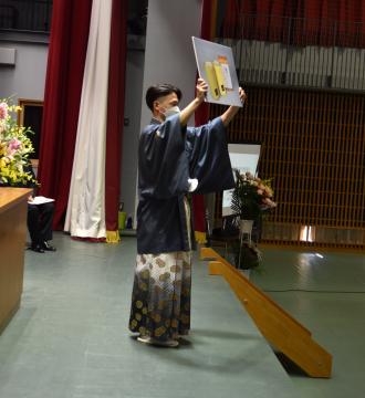 ステージ上で記念品を両手に持ち頭上まで掲げている羽織袴姿の男性の写真