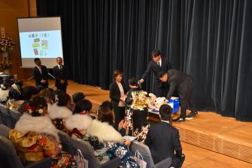 振袖やスーツ姿の参加者が着席している前方で関係者が抽選会をおこなっている様子の写真