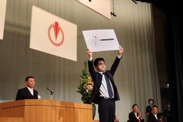 町長が立つ演台の前方で記念品のボードを両手に持ち頭上に掲げているスーツ姿の男性の写真