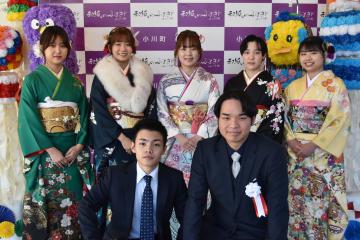 後方に小川町のバックボードそして両脇に吹き流しが飾ってある場所で前列にスーツ姿の男性2名が座り後列に振袖姿の女性5名が立っている記念撮影の写真