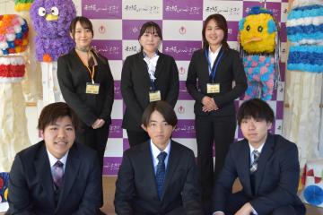 後方に小川町のバックボードそして両脇に吹き流しが飾ってある場所で前列にスーツ姿の男性3名が座り後列にスーツ姿の女性3名が立っている記念撮影の写真