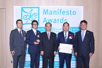マニフェスト大賞のバックボードの前でトロフィーや賞状を持った男性2名と関係者3名が記念撮影を行っている写真