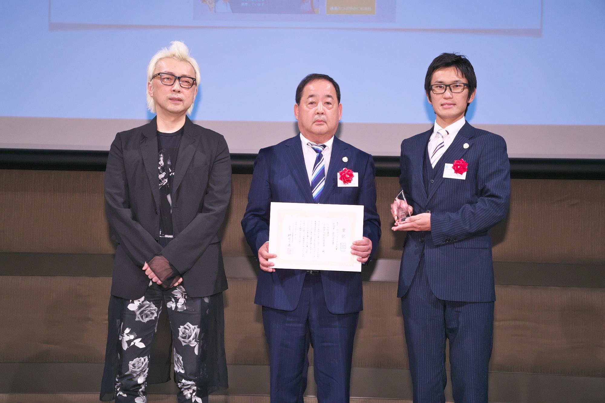 賞状を持った男性とトロフィーを持った男性そして箭内道彦氏3名で横一列に並び記念撮影をしている写真