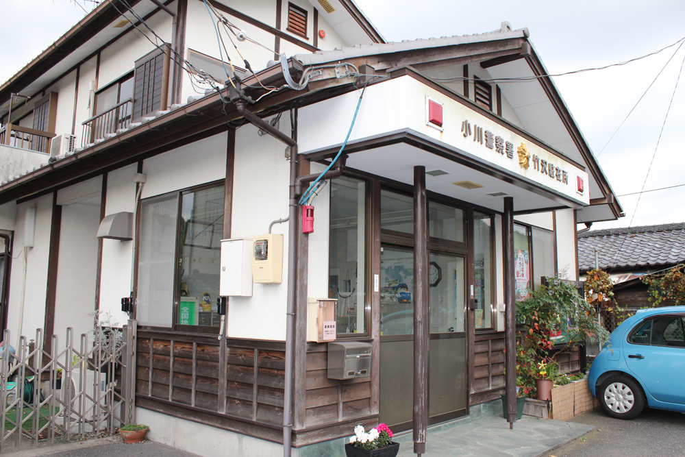 入口前の花壇とプランターには花が咲いていて2階和風造り外観の小川警察署竹沢駐在所をななめ前方から撮影した写真