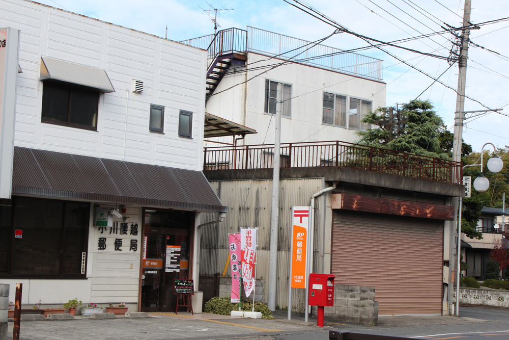 茶色のシャッターが閉められている建物の隣に建つ白い外観で黒い庇がかかる小川腰越郵便局をななめ前方から撮影した写真
