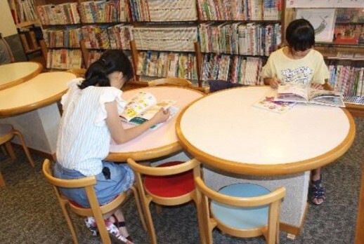 図書館内に設置された丸いテーブルの席で本を読んでいる2名の女の子の写真