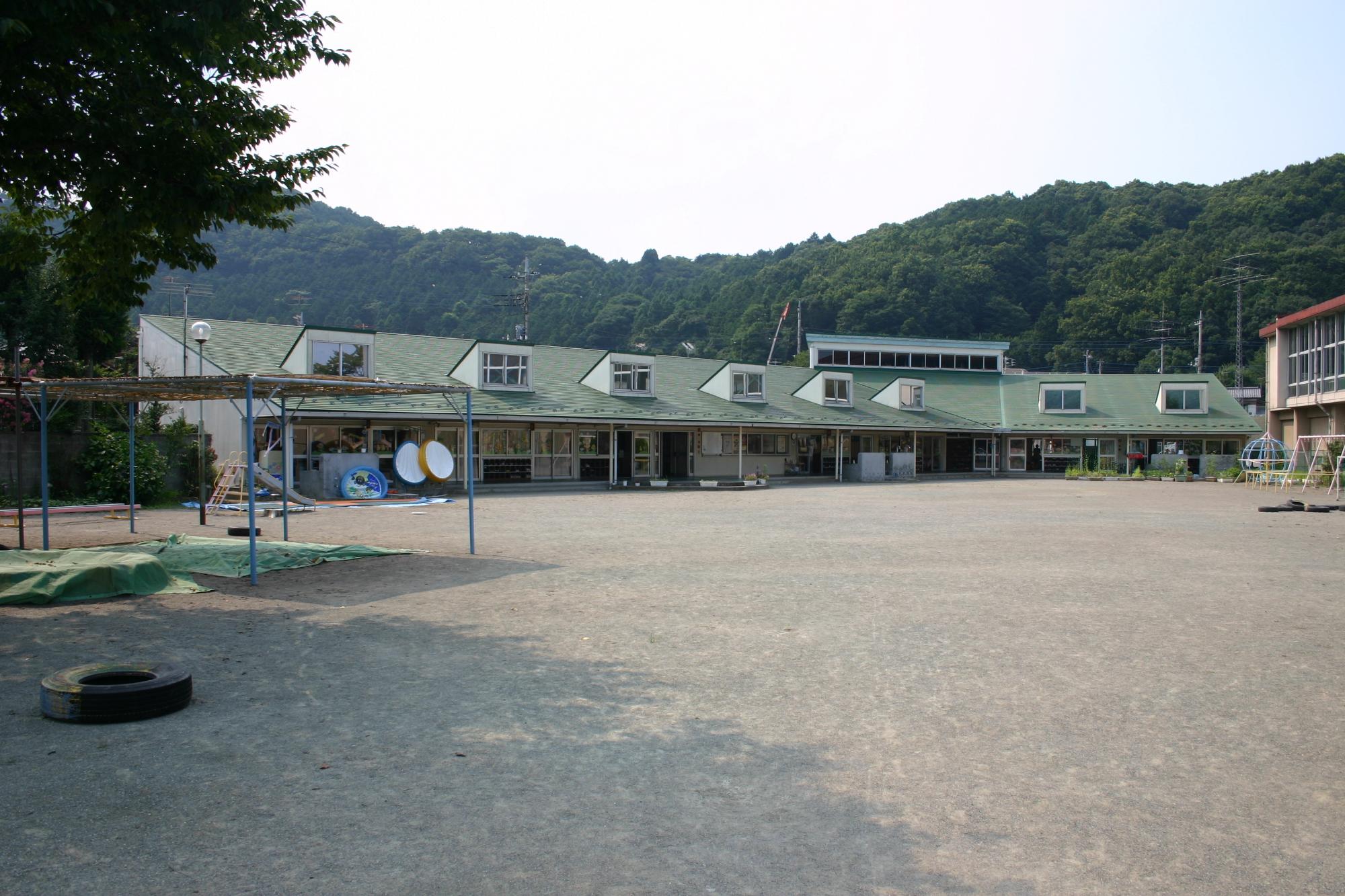 広々とした園庭の奥に建つ薄緑色の屋根の保育園の建物外観の写真
