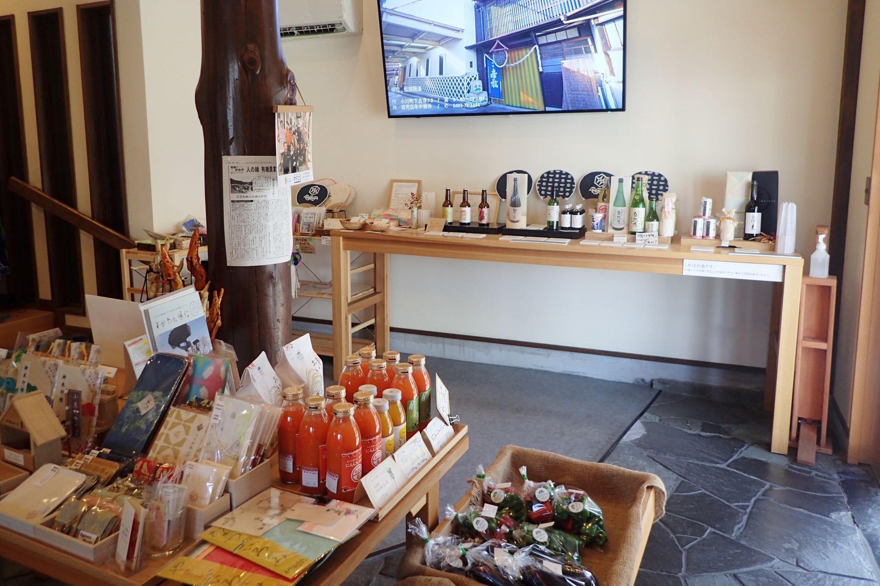 酒蔵の映像が映し出されたモニターが壁に設置された室内に、お酒や野菜に特産品などの沢山の商品が陳列されているむすびめの写真