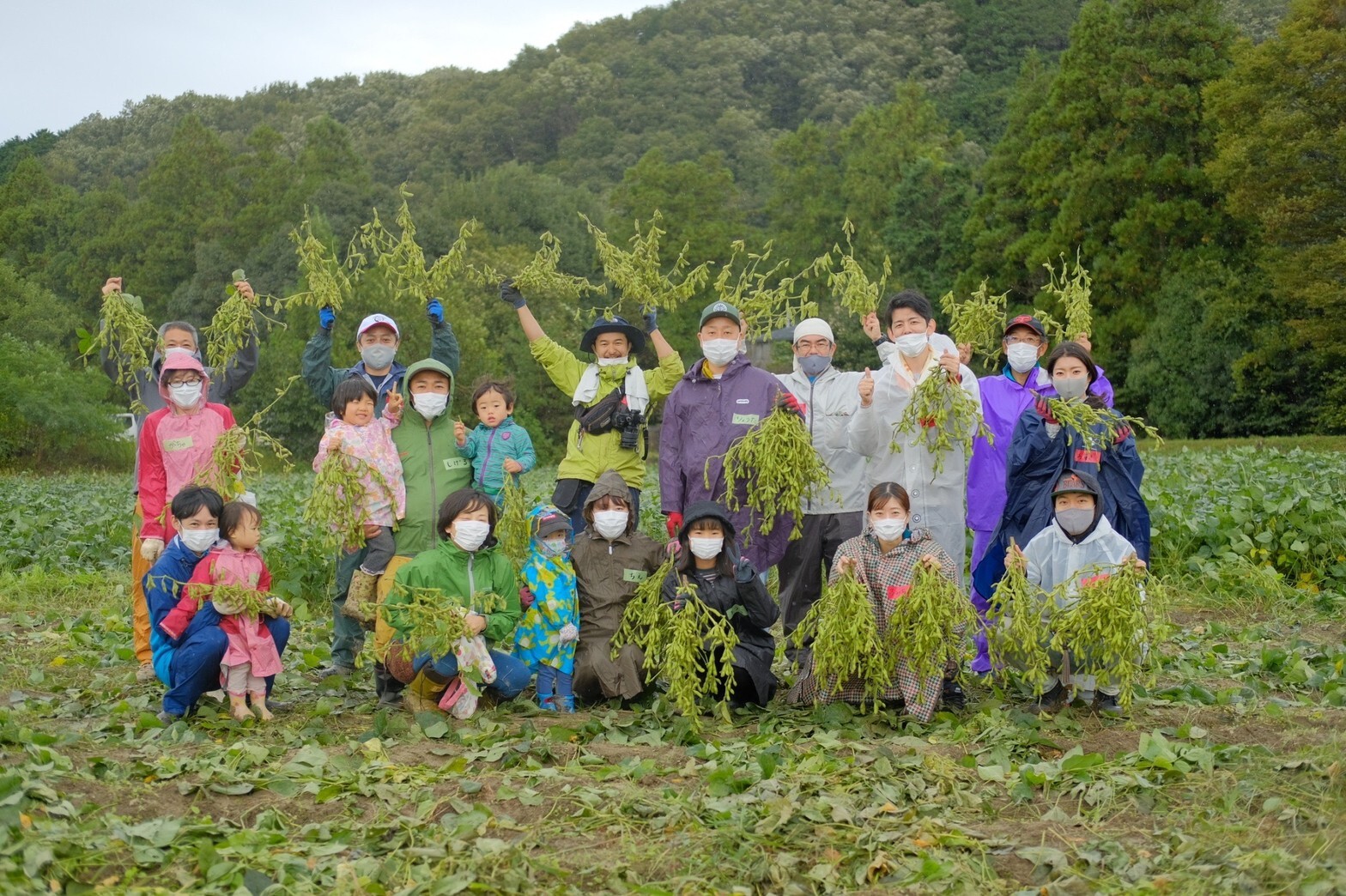 帽子を被りレインコートを着た参加者が収穫した枝豆を束ごと持っている集合写真