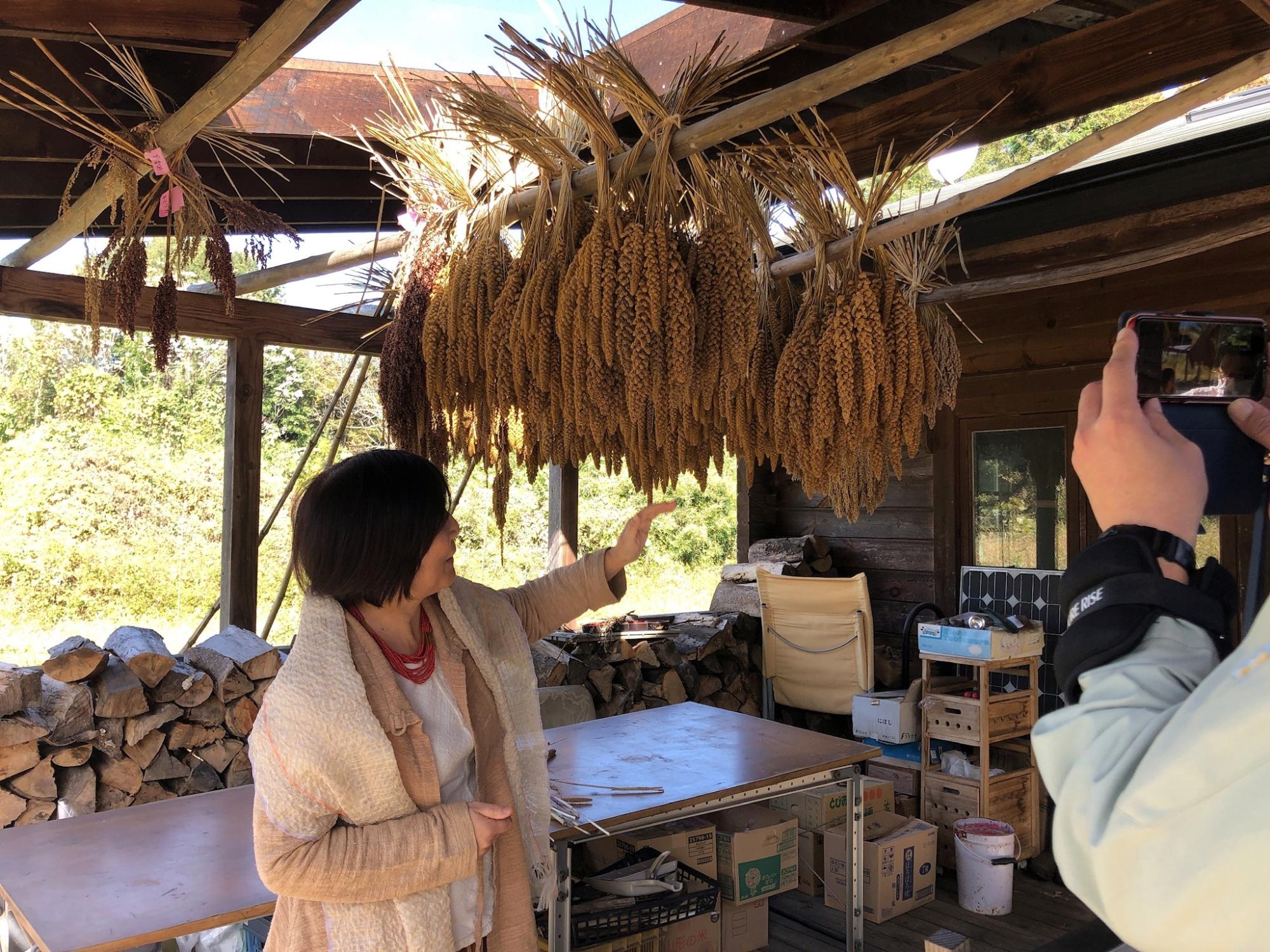 軒先に掛けられた植物を笑顔で触れている女性の様子を右側に立つ男性が撮影している写真