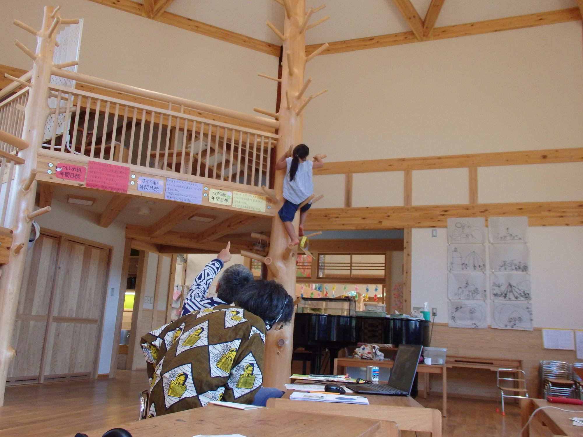 枝のついた木の様な柱に女の子が登っている様子を男性が指さし、もうひとりの男性がノートパソコンを広げて見ている写真