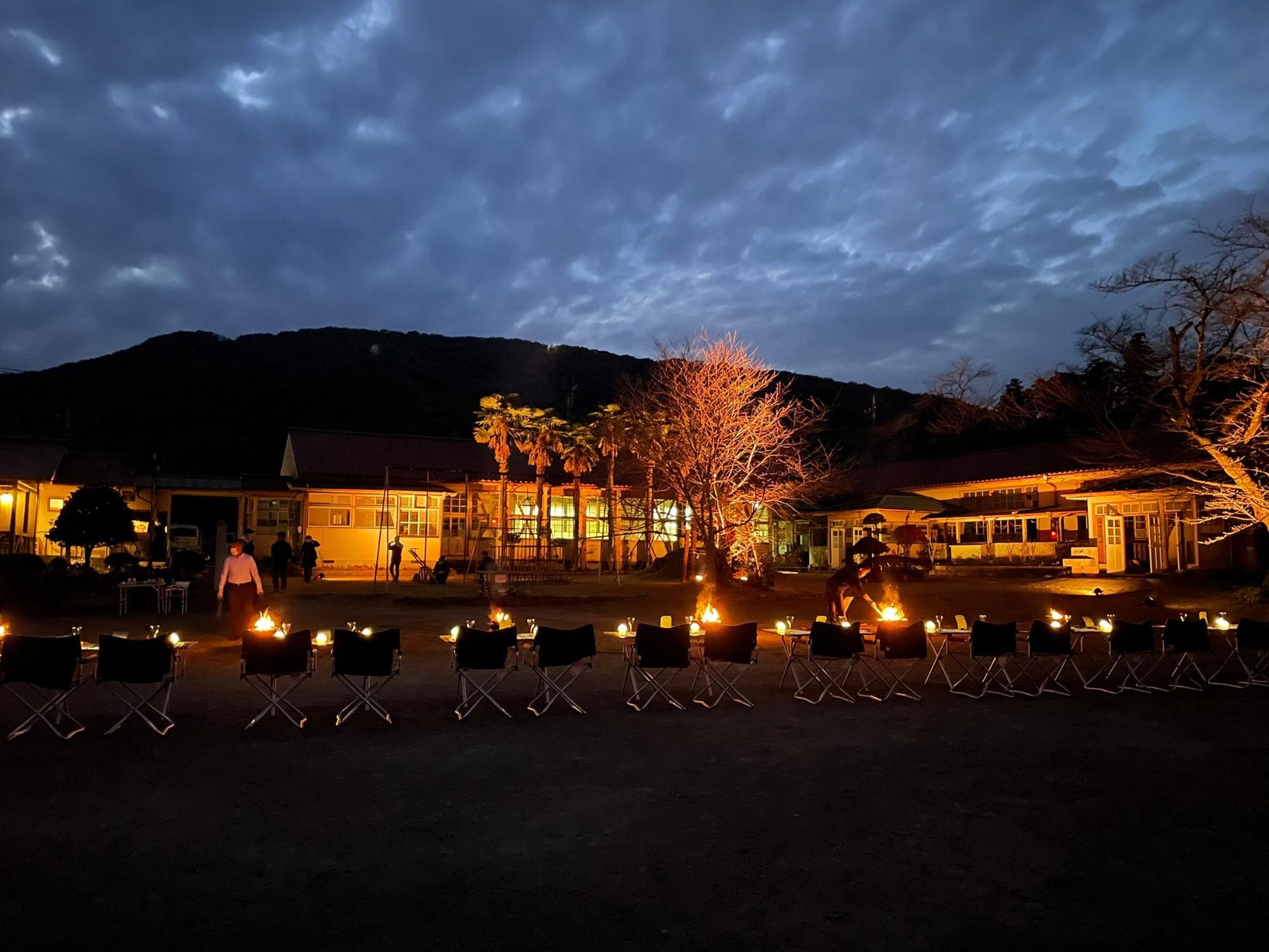 校庭に2脚ずつセットされた簡易テーブルが横一列に並んで設置され、旧下里分校の校舎に灯りが灯された写真