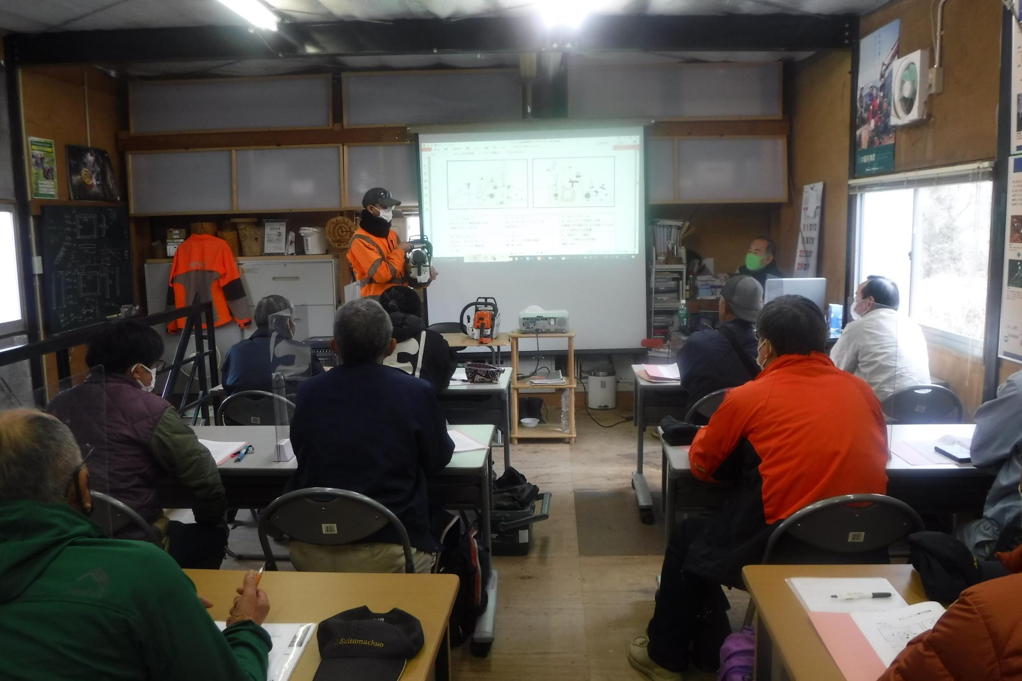 長机が並んだ室内前方に資料が映し出されたスクリーンが設置され、左側に立つ男性講師の話を参加者が熱心に聞いている安全講習の様子の写真