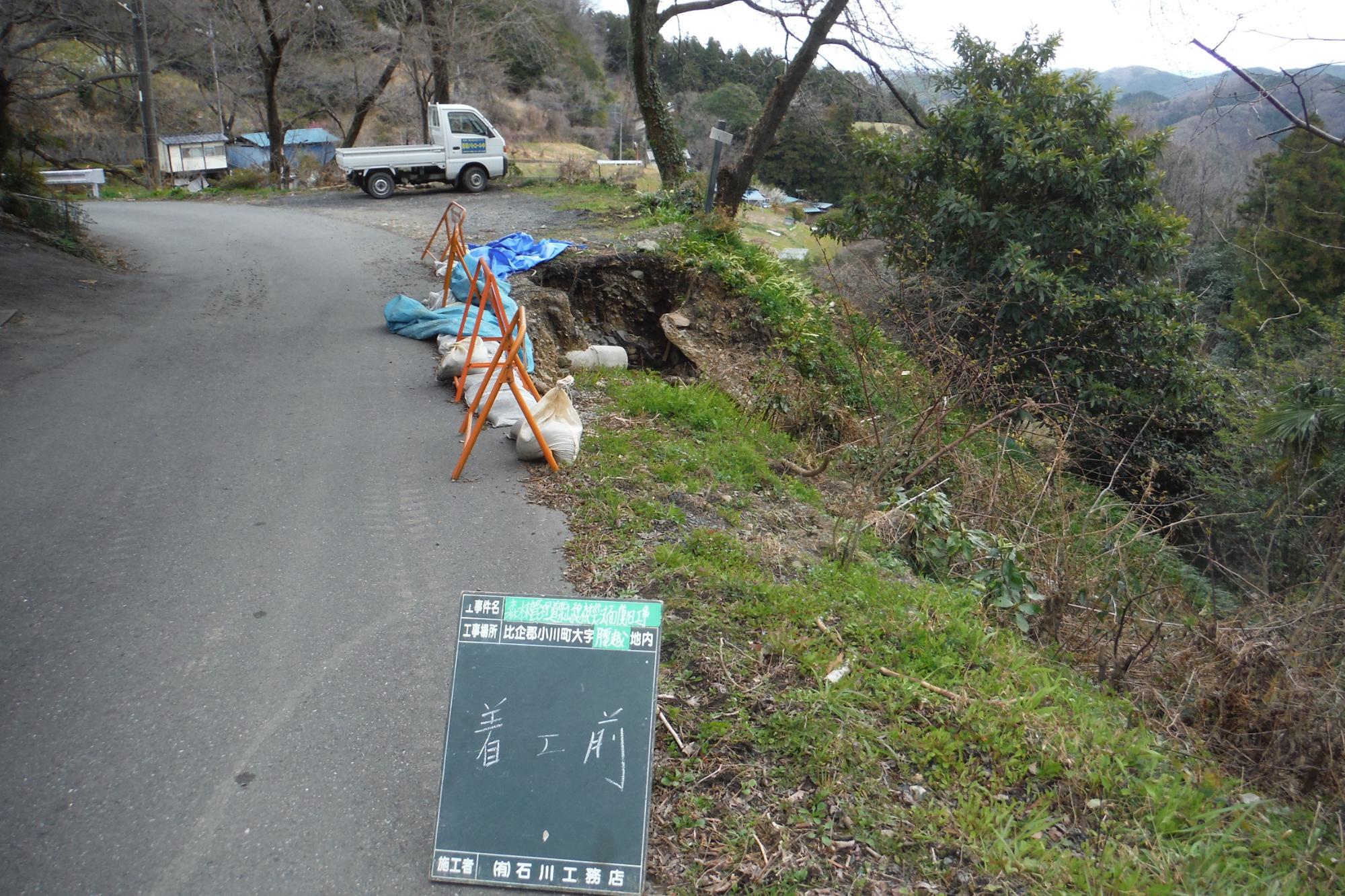 「着工前」と記載された工事用黒板の奥の右側の道路と斜面の境目に4つのオレンジ色のバリケードが設置され、斜めに木が立っている手前の斜面が崩れている復旧工事前の写真