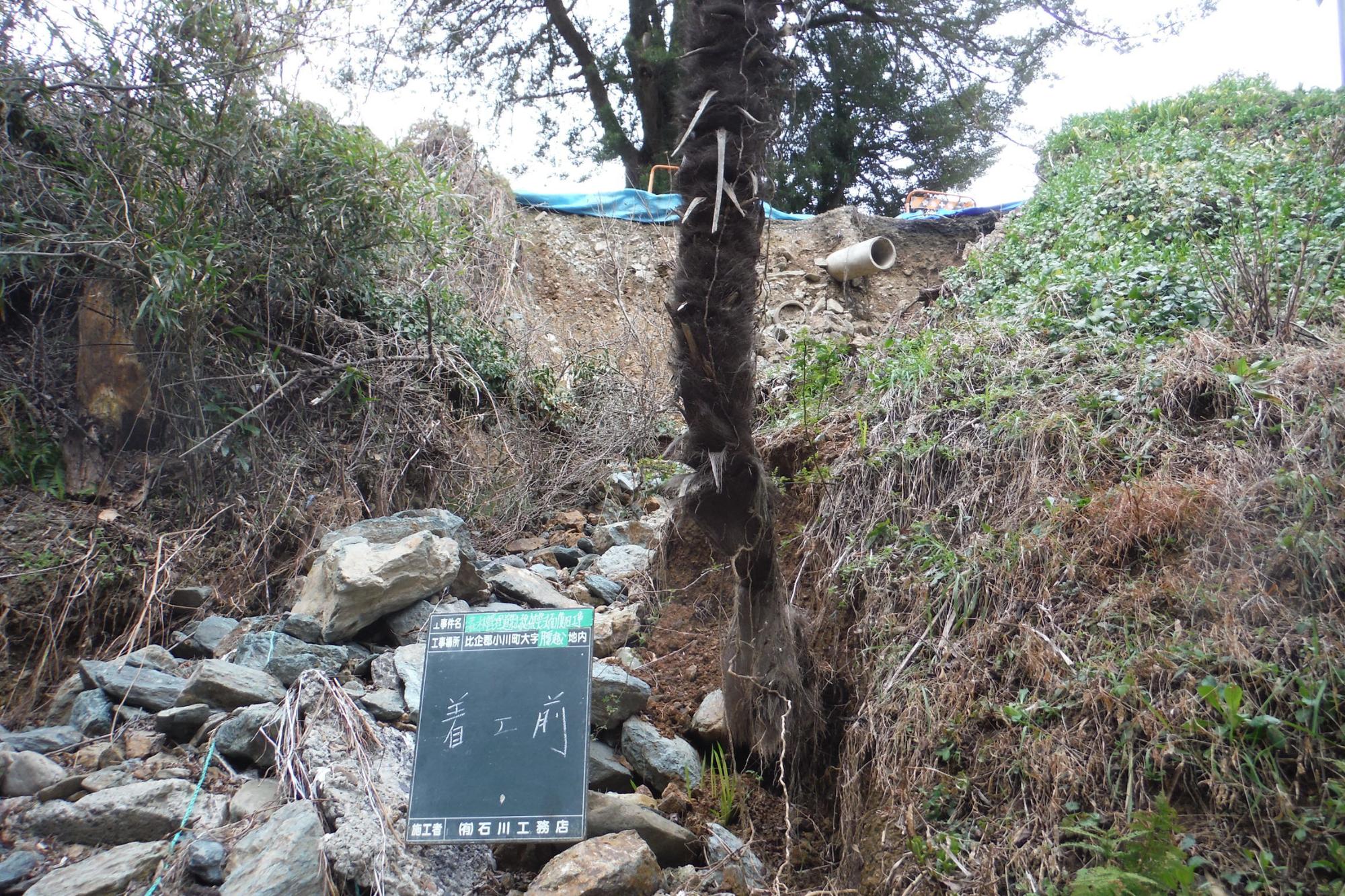災害で大きな石が扇状に流出し一本の木が立つ左側に「着工前」と記載された工事用黒板が設置され、流れてきた高所にビニールシートが敷かれている復旧工事前の写真