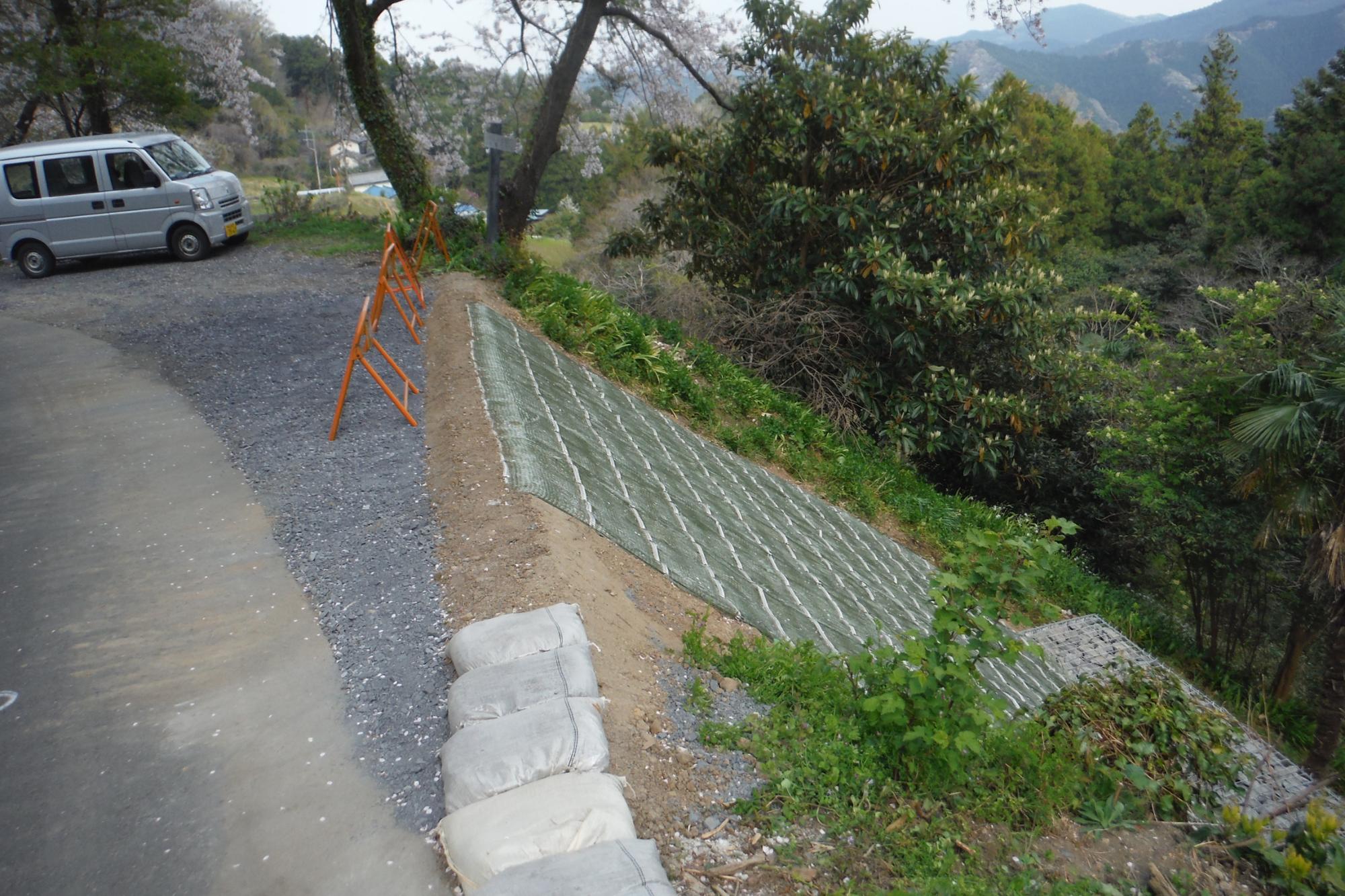 道路と斜面の境目に4つのオレンジ色のバリケードが設置され、崩れた斜面の場所に緑色のシートが設置された復旧工事後の写真