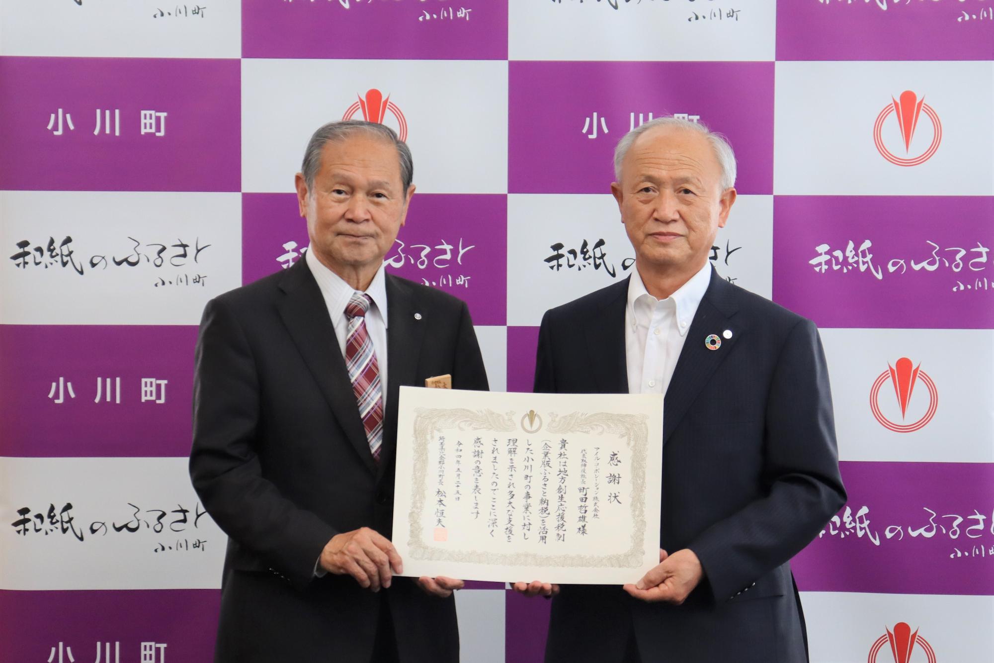 小川町のバックパネルの前で、アイル・コーポレーションの町田社長と松本町長が一緒に感謝状を持っている贈呈式の様子の写真