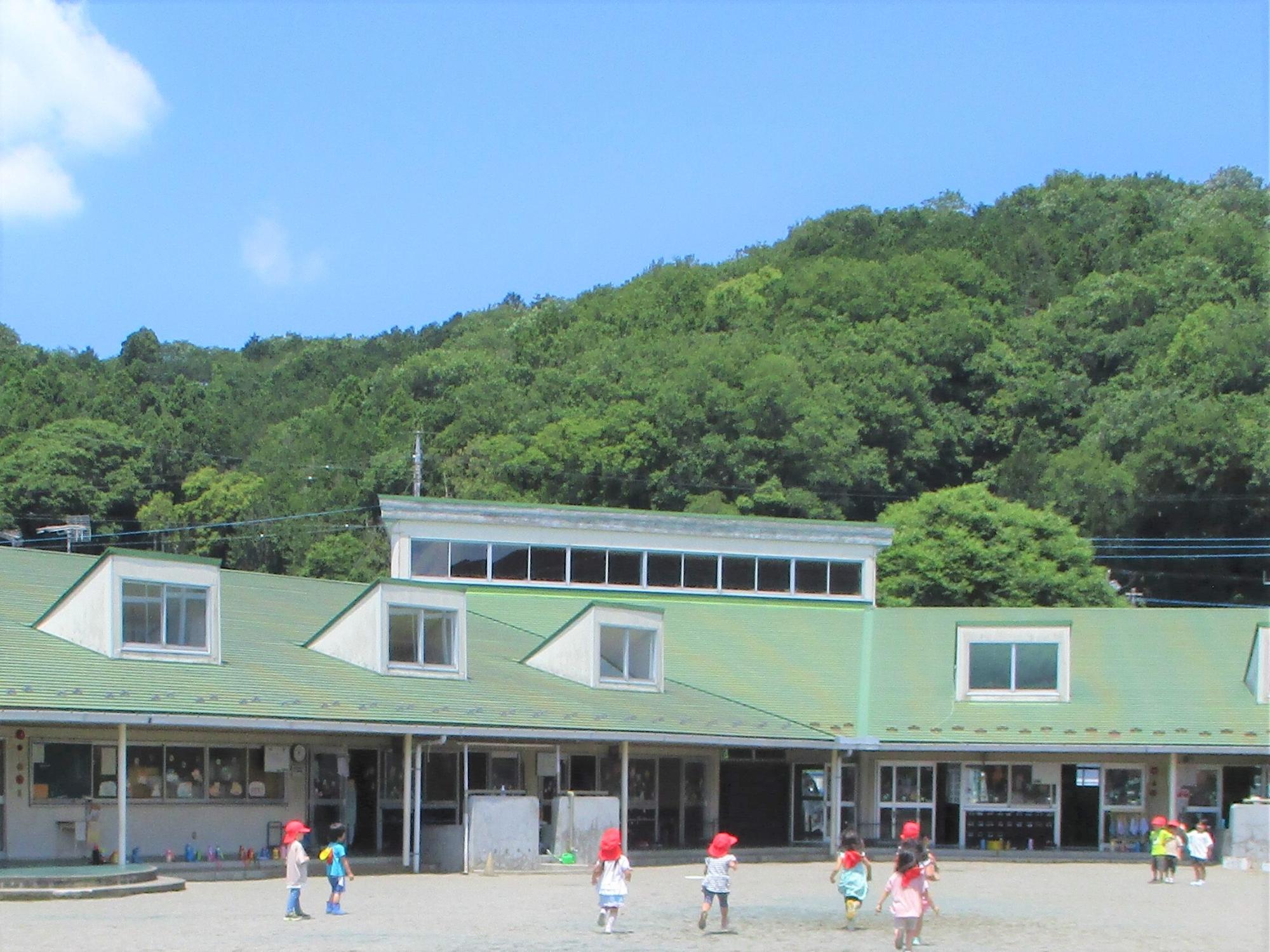 園児たちが元気いっぱいに遊んでいる園庭の奥に、小さな山の手前に建つ薄緑色の屋根の保育園の写真