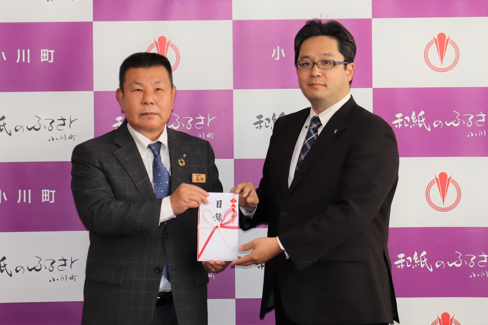 小川町のバックパネルの前で、東武トップツアーズの竜江取締役と島田町長が一緒に目録を持っている贈呈式の様子の写真