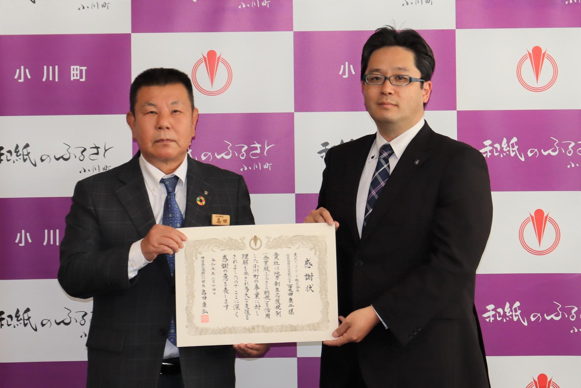 小川町のバックパネルの前で、東武トップツアーズの竜江取締役と島田町長が一緒に感謝状を持っている記念写真