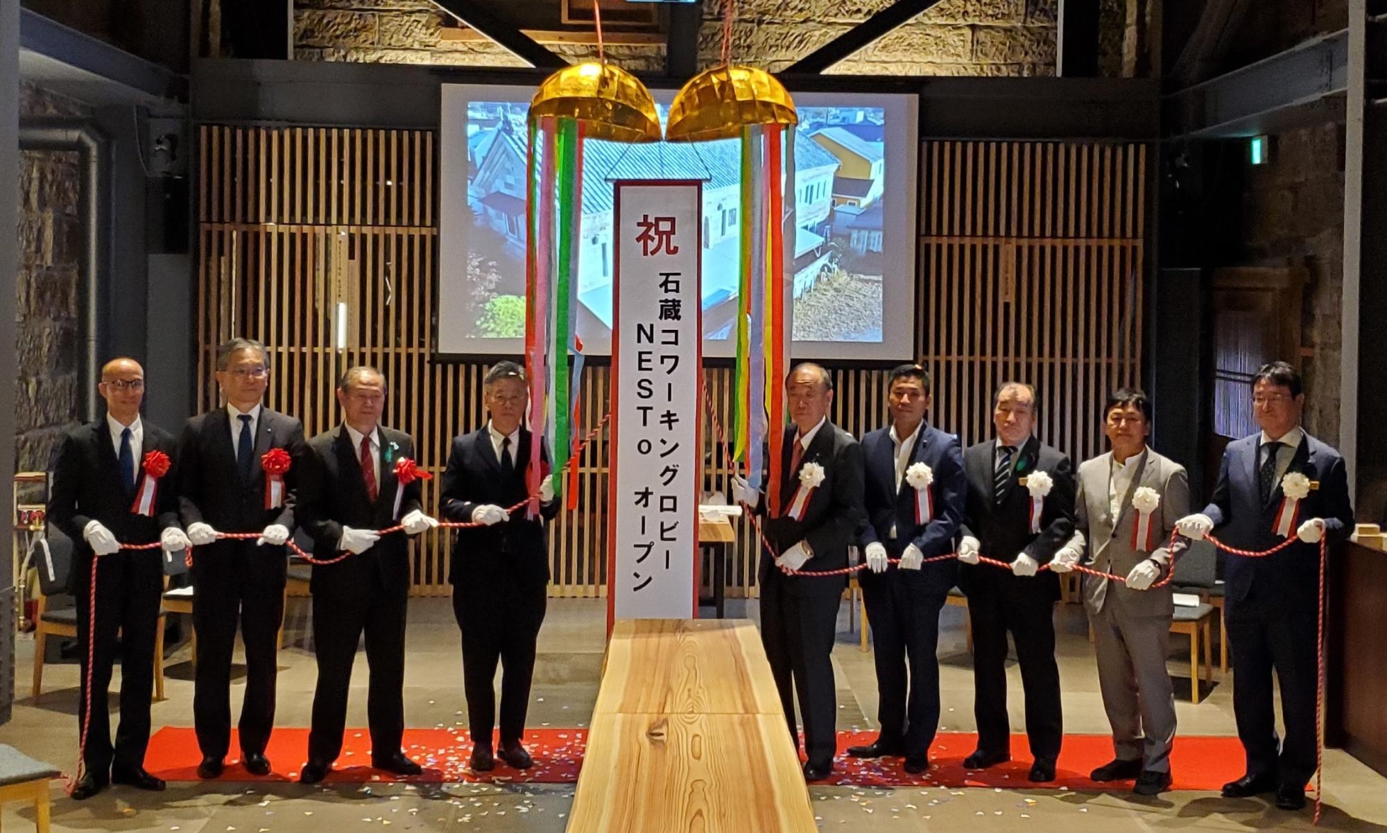 くす玉が割られた中から「祝 石蔵コワーキングロビーNESToオープン」と書かれた幕が現れ、9名の関係者がテープカットを行おうとしているオープニングセレモニーの様子の写真