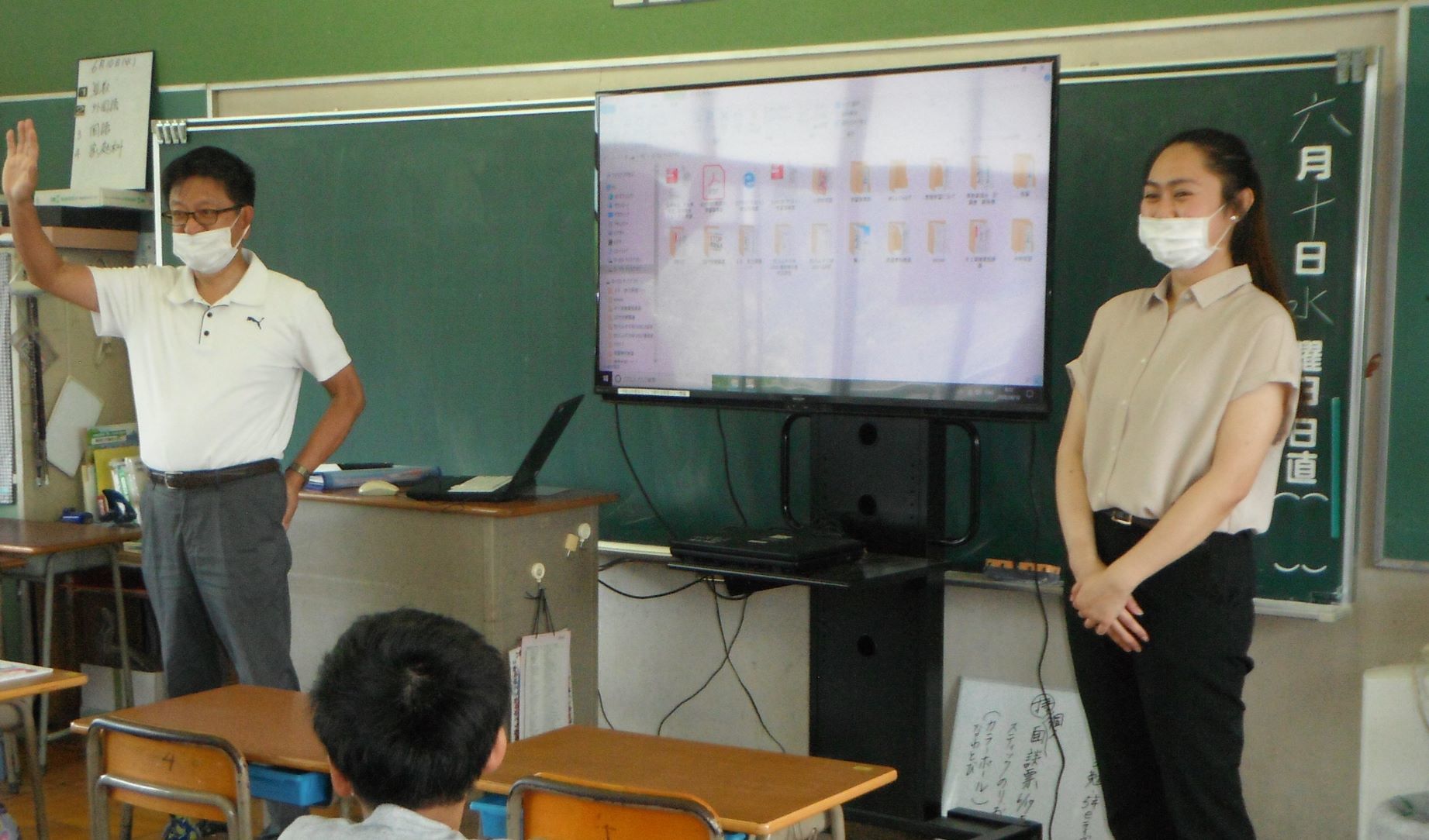 黒板の前に大型モニターが設置され、左右に男性教師と女性が立っている授業の様子の写真