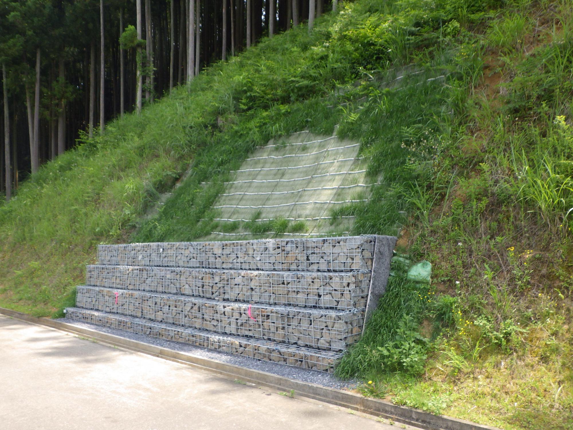 細い木が立ち並ぶ手前の崩れた斜面に緑色のシートが被され、銀色の柵で囲われた石塀が設置された復旧工事後の写真