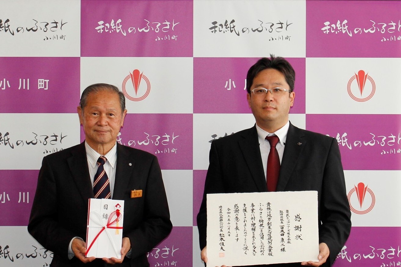 小川町のバックパネルの前で、松本町長が両手で目録を持ち、東武トップツアーズの竜江取締役が両手で感謝状を持ち並んでいる記念写真