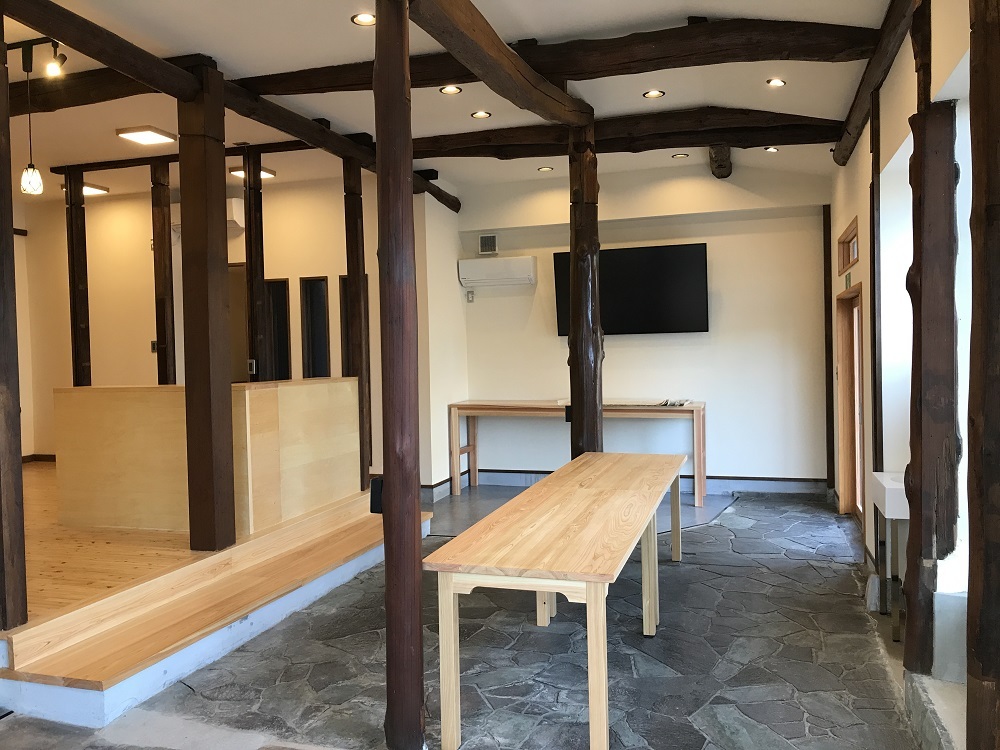 室内全体に茶色の木製の柱が設置され、左側に温かみのあるフローリング床と木製の家具、右側奥の壁に大きなモニターがあり中央に木製の長テーブルが設置されたむすびめ内観の写真