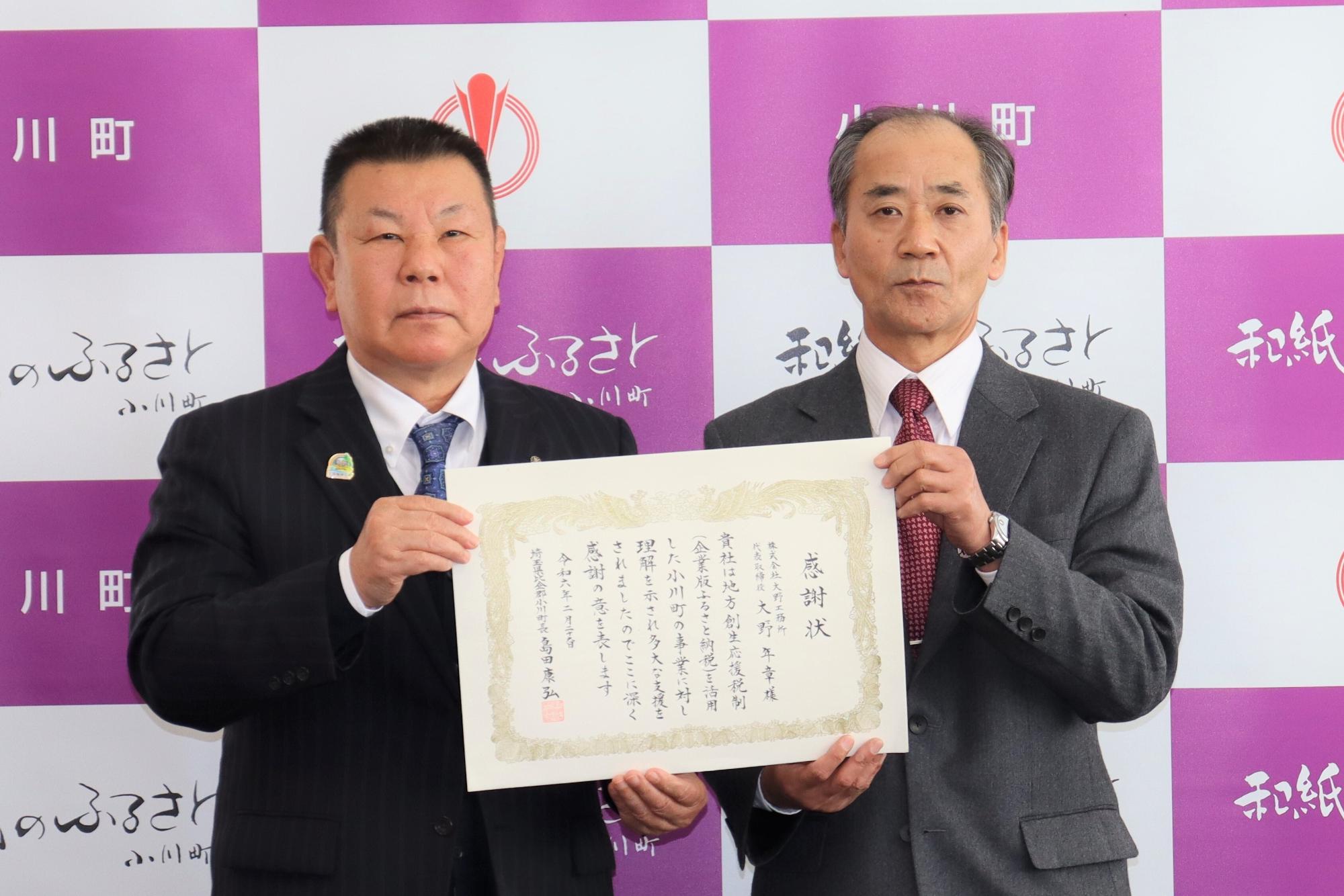 小川町のバックパネルの前で、大野工務所代表の男性と町長が一緒に感謝状を持っている贈呈式の様子の写真