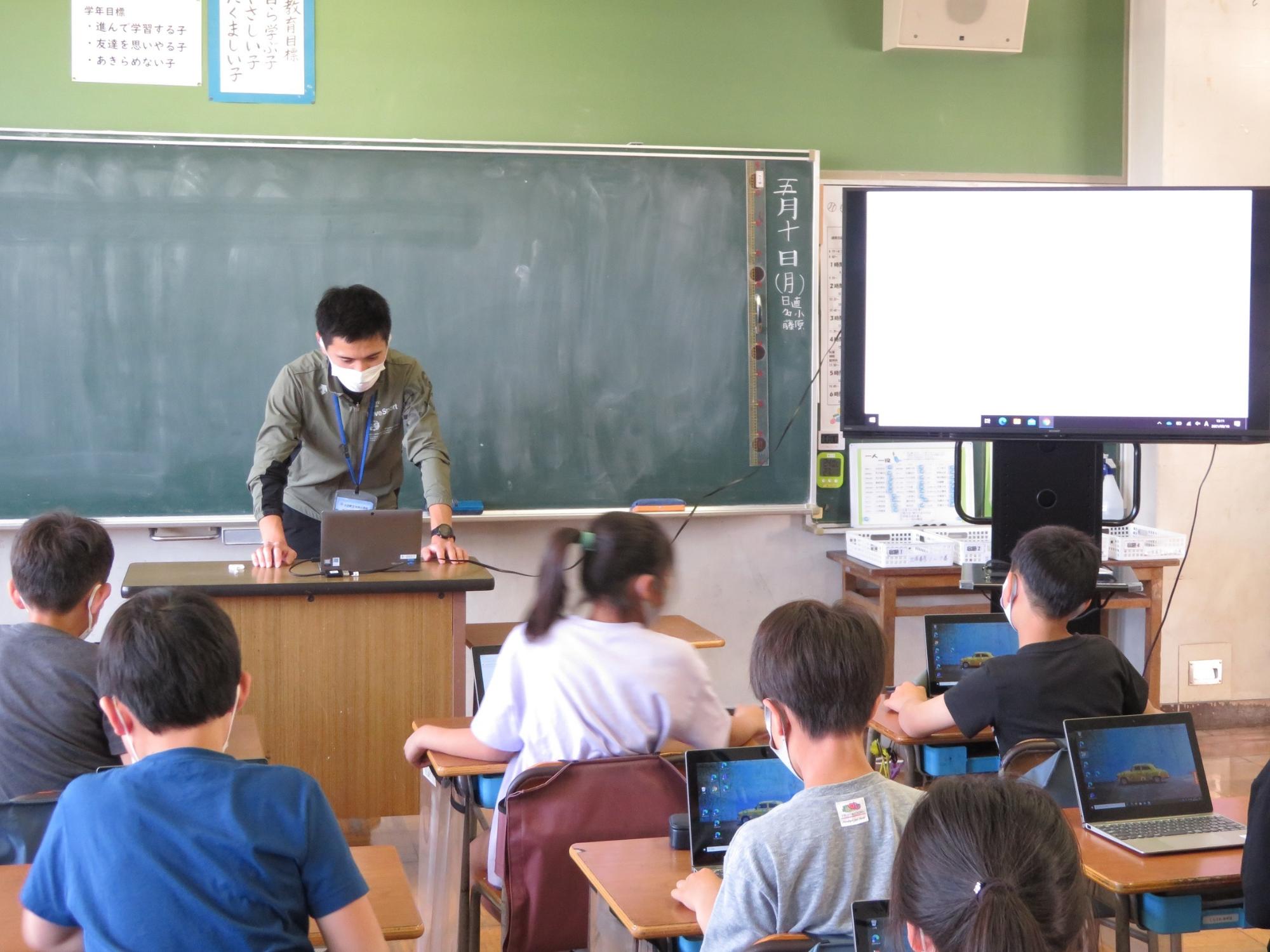 大きなモニターが黒板の右側に設置された教室で、児童たちの机の上にそれぞれノートパソコンが置かれ授業が行われている様子の写真