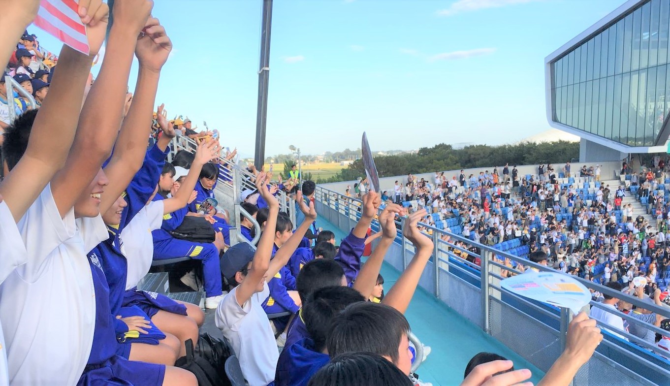 青色のジャージ姿の生徒たちが観客席で両手を上げて観戦している様子の写真