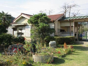 樹木や植木にところどころ花が咲いている庭の奥に建つ、赤色の屋根の整備前の用務員棟の建物外観の写真