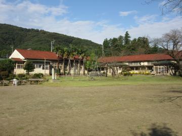 山の麓の敷地内に校庭とL字型に建つ赤色の屋根の校舎棟全体を撮影した写真