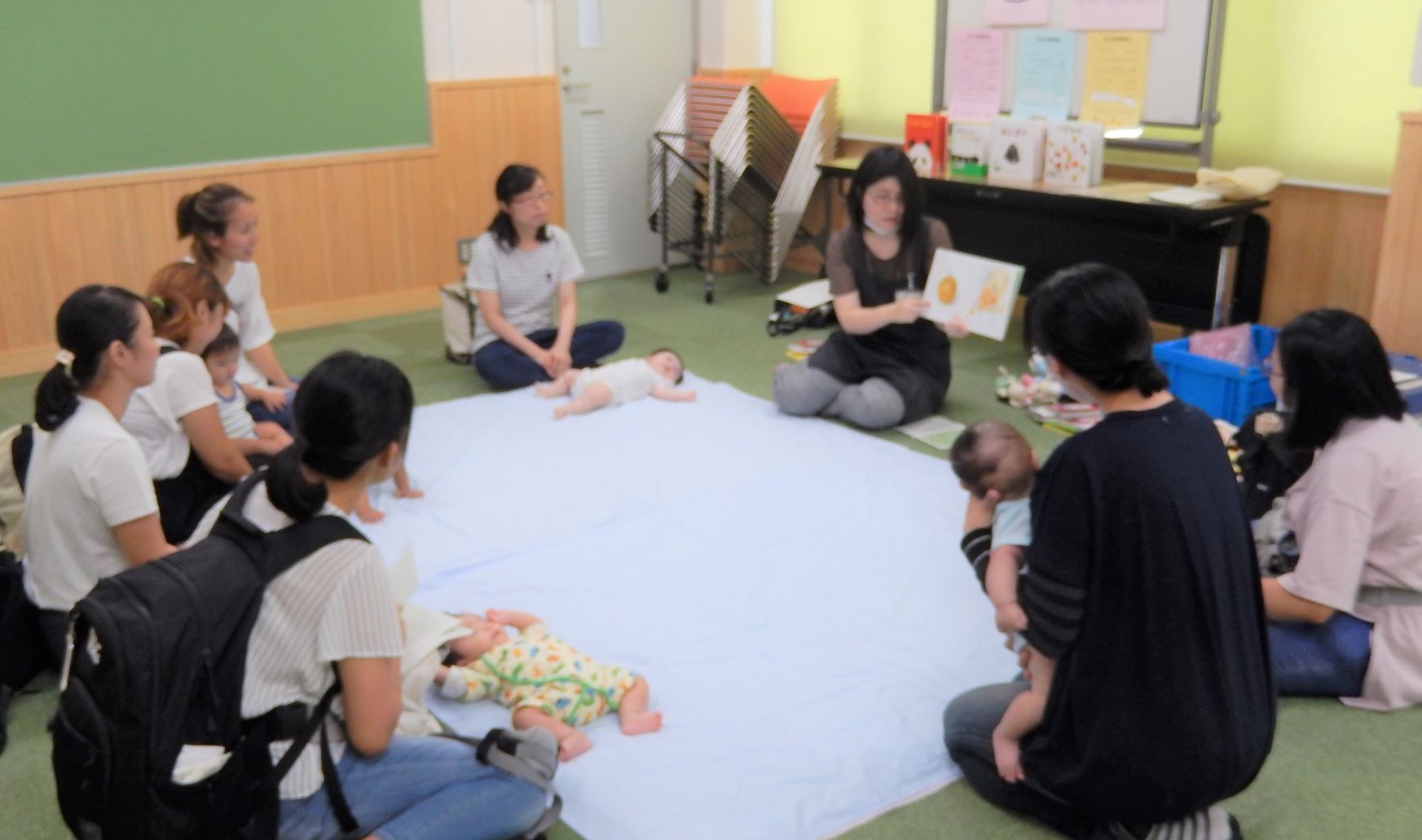 赤ちゃんを寝かせた大きなタオルケットを囲んで参加した母親が座り、読み聞かせを行っている女性の話を聞いている親子の参加者の写真