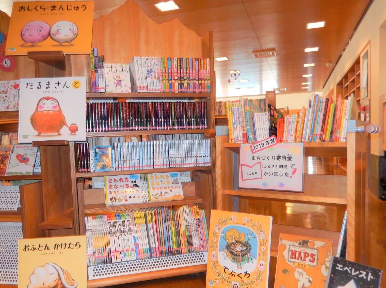 「2019年度まちづくり寄付金（ふるさと納税）でかいました！」と書かれたポップが沢山の本が並んだ木製の本棚の上に貼られている写真