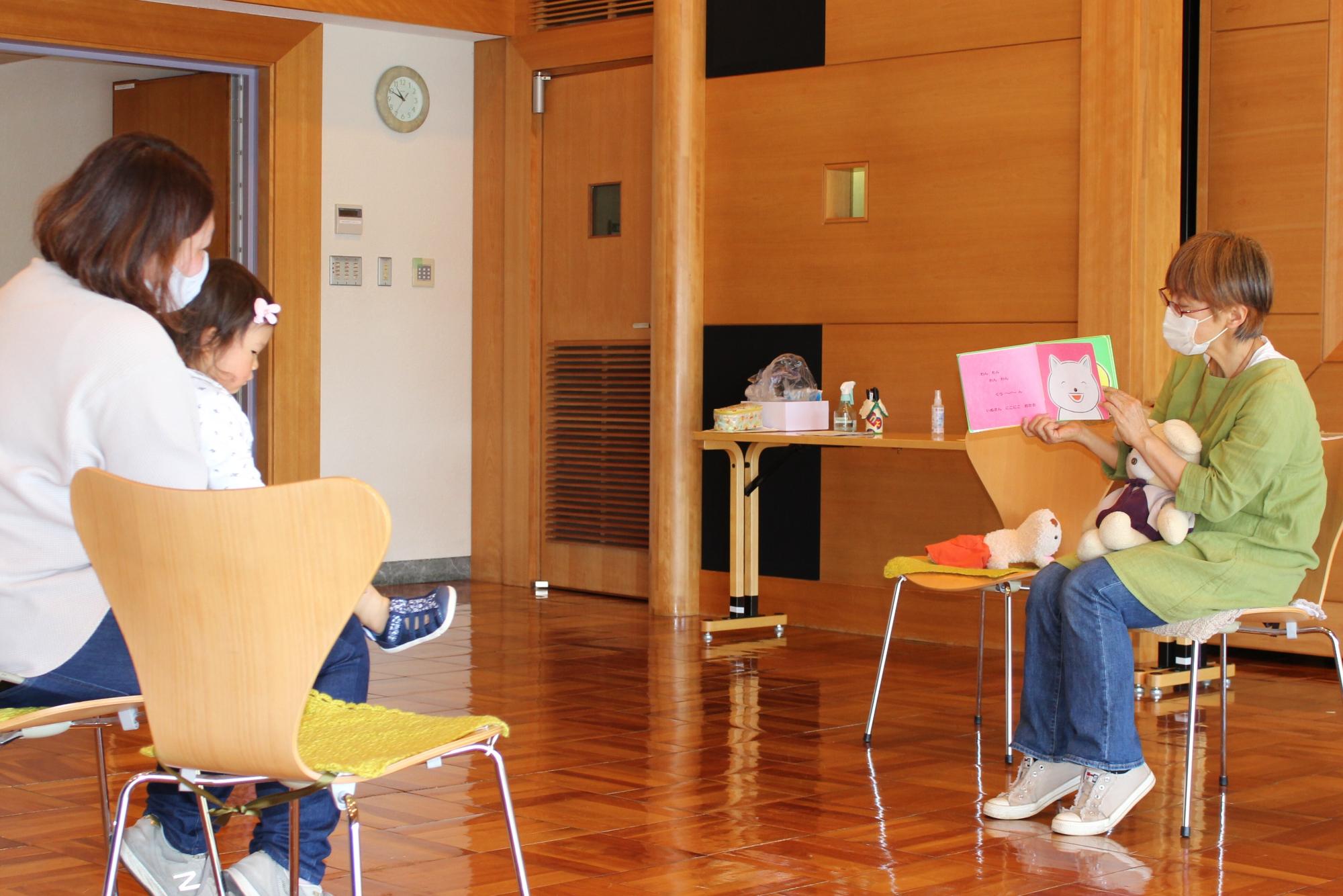 椅子に座って絵本を持っている黄緑色の服を着た女性と向かい合って座っている母親と女の子が話を聞いている読み聞かせの様子の写真