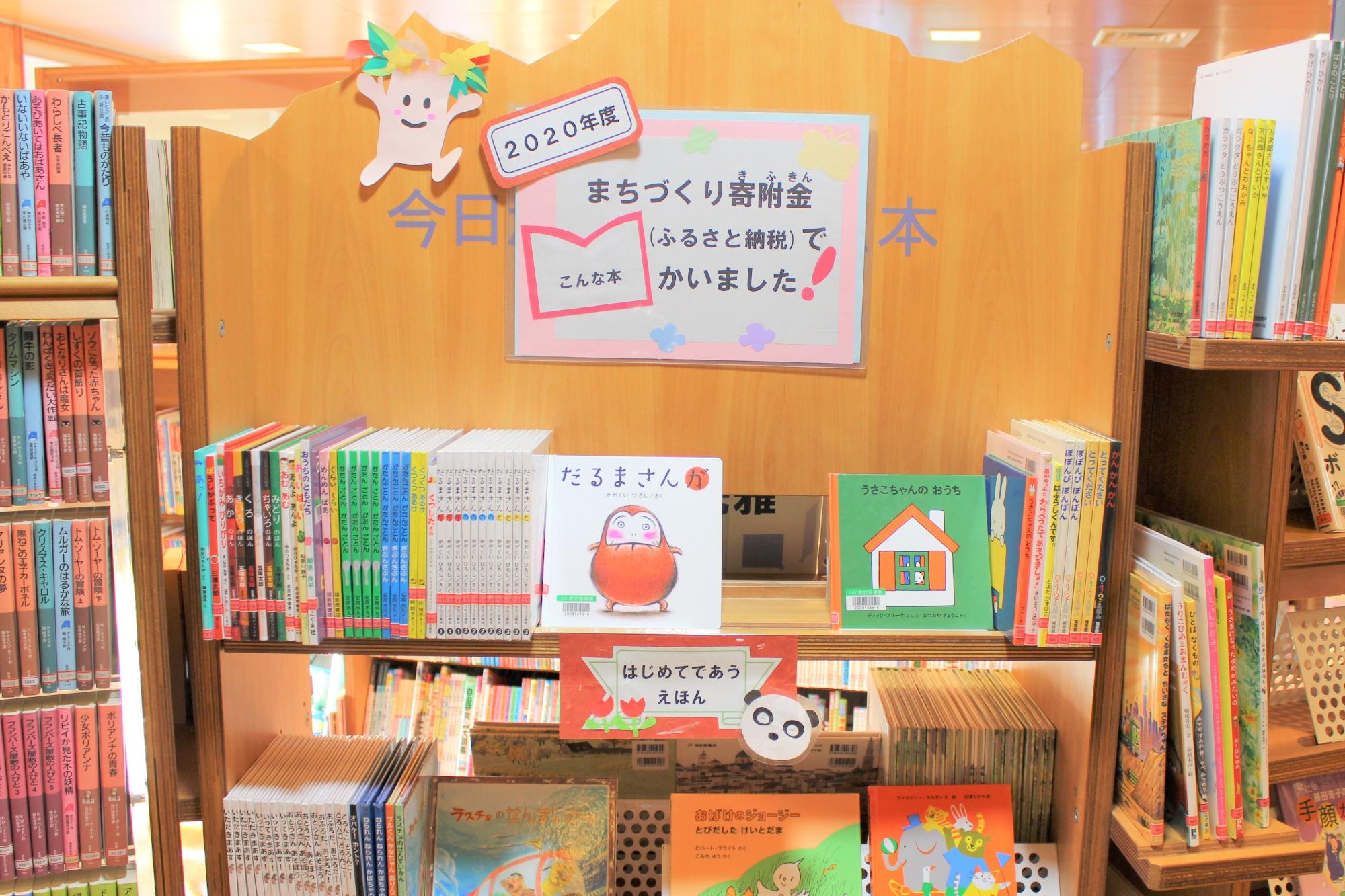 「2020年度まちづくり寄付金（ふるさと納税）でかいました！」と書かれたポップが沢山の本が並んだ木製の本棚の上に貼られている写真