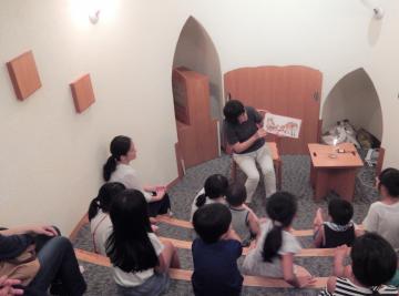 椅子に座った女性が絵本を見ており、その前には子供たちが座っている読み聞かせ風景の写真