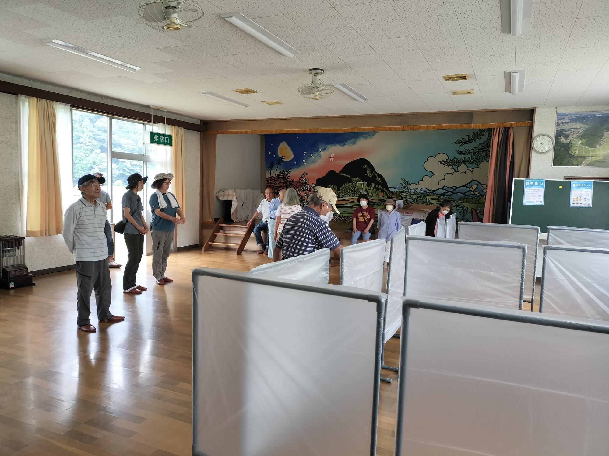 小川町農村センターのフローリングの床に男性女性の参加者の方々がパーテーションを立て区画を作っている様子を撮影した写真