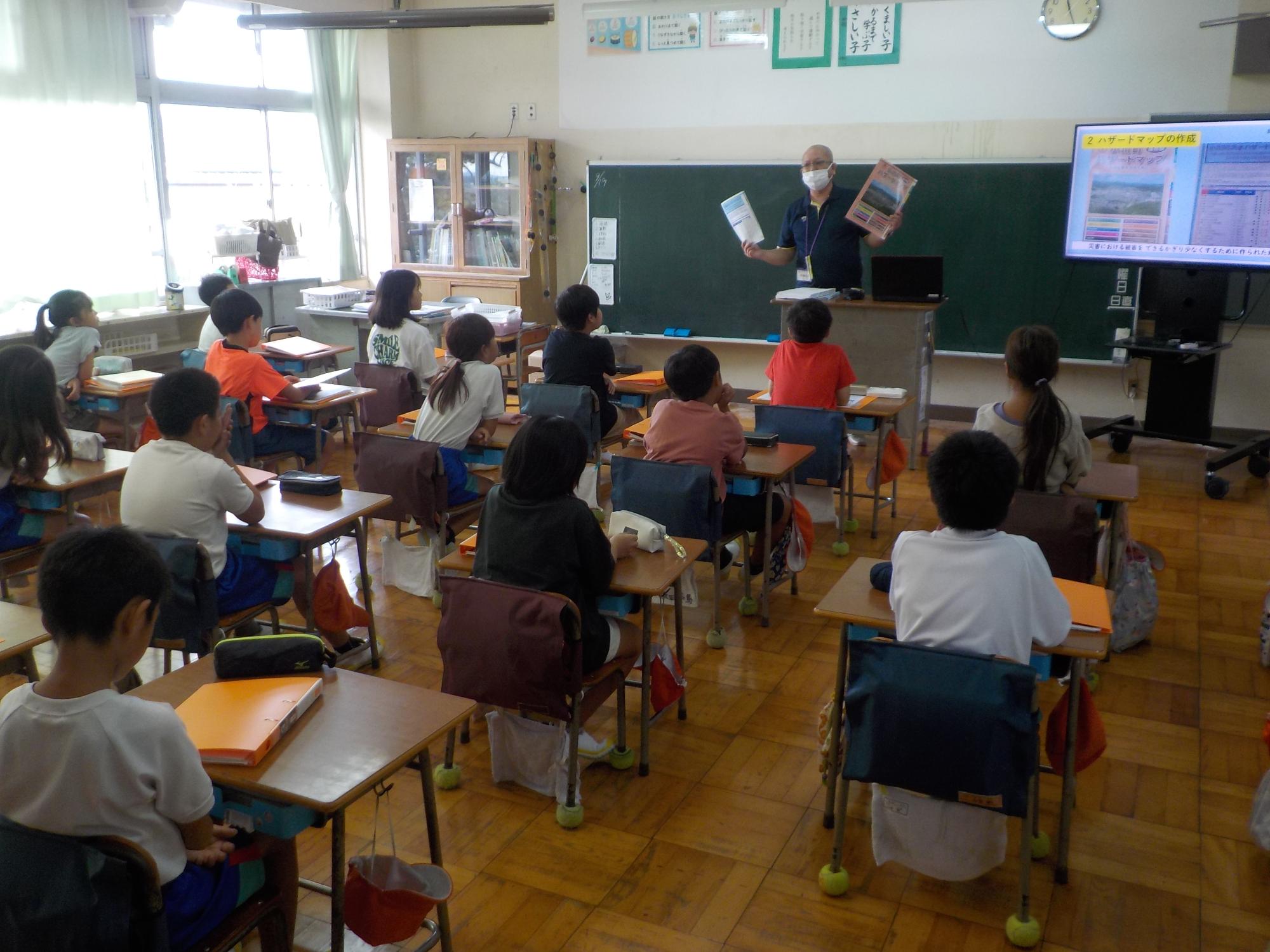 八和田小学校の教室の黒板の前にハザードなど2枚を手に持つ講師の男性の説明を机に座り真剣に聞いている生徒達を後方から写した写真