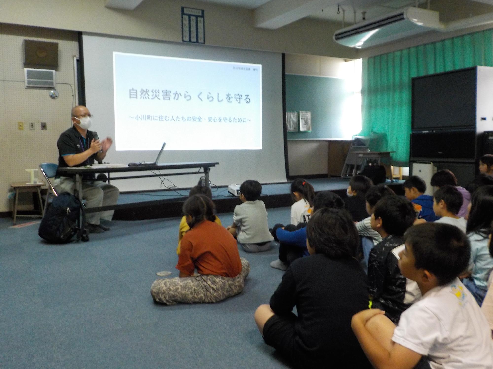 スクリーンの横に講師の男性が座り、子供たちが視聴覚室で座りながら説明を聞いている写真