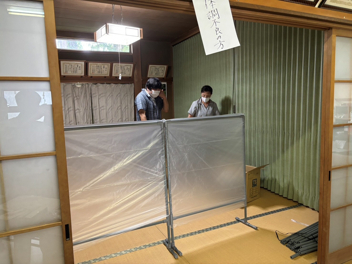 諏訪会館の和室の部屋で鴨居に「体調不良の方」と書かれた紙が張られ、パーテーションの奥に立ち区画割を行う参加者2人を撮影した写真